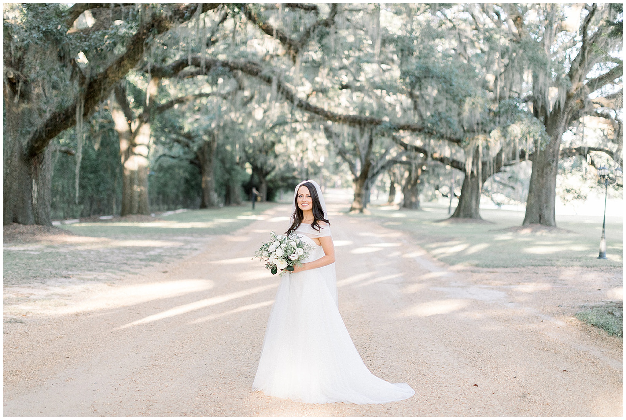 A bride in a white tule dress holds her white flower bouquet on an unpaved road lined with oak trees covered in moss