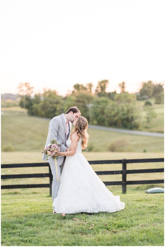 Bride and groom smile and hold hands while walking across the open countryside at their elegant barn wedding.