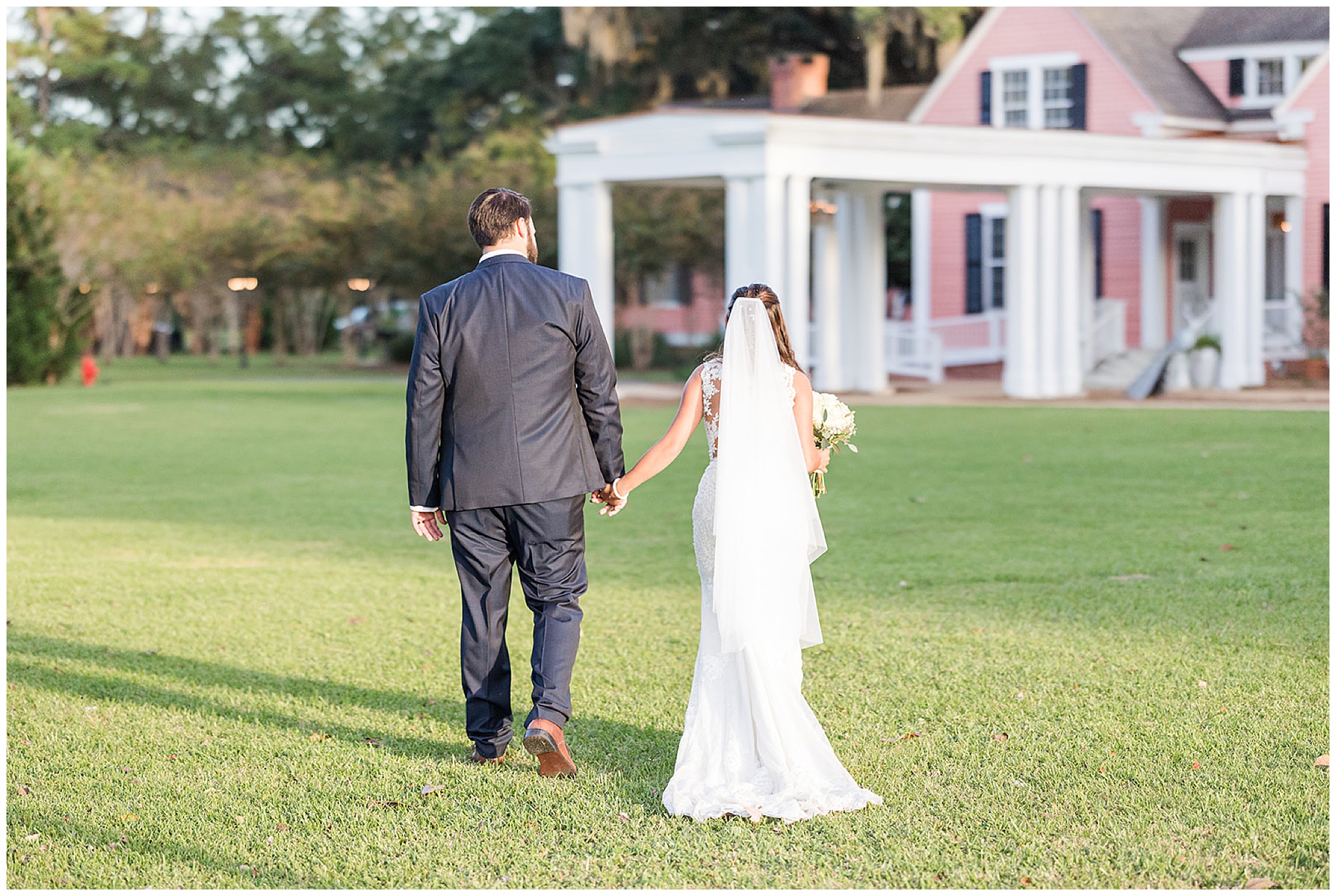 Newlyweds hold hands while walking through a manicured lawn towards a pink building