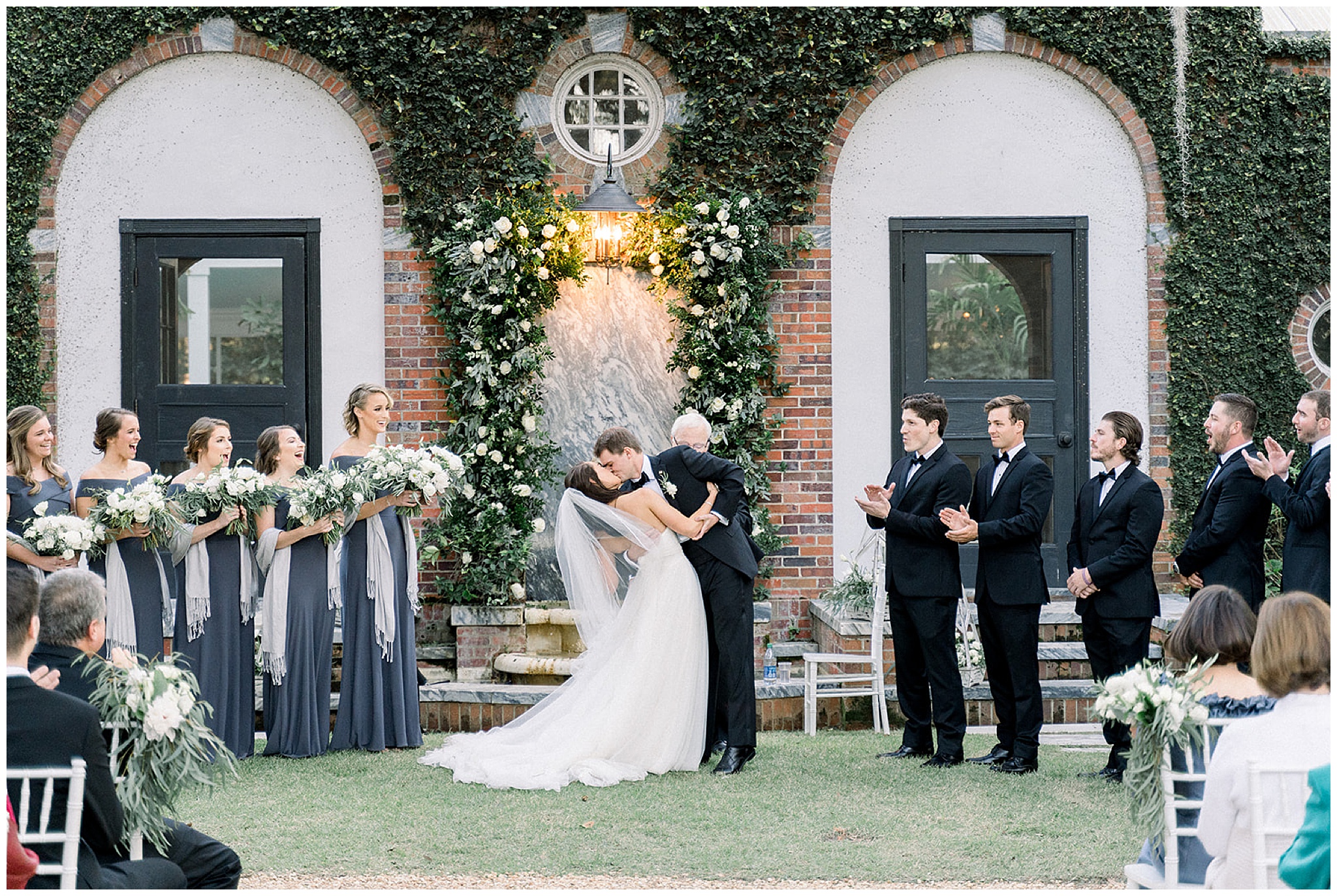 Newlyweds kiss for the first time as their wedding party and guests cheer in front of an ivy-covered brick wall with archways for doors